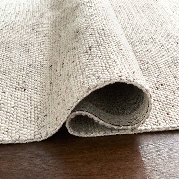 Close-up of rolled neutral woven area rug showing chunky loop texture and edge binding on dark wood floor
