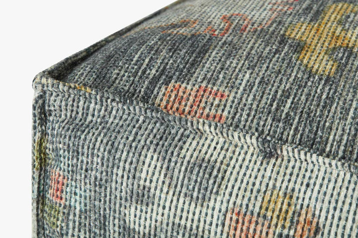 Close-up of patterned upholstered ottoman corner showing blue-gray woven fabric with multicolor motifs and stitched edge detail