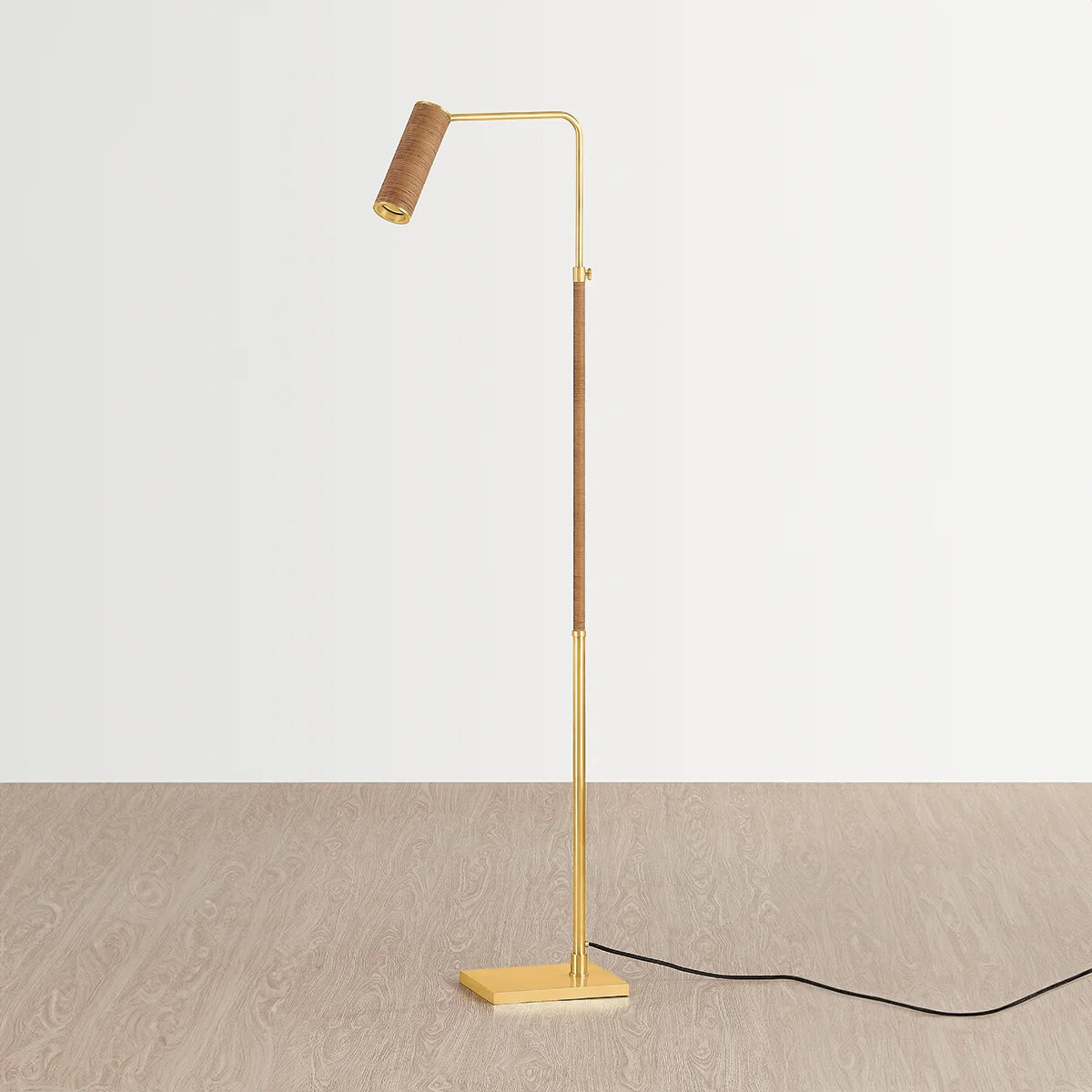 Brass floor reading lamp with woven rattan-wrapped stem and adjustable spotlight head on square base, shown on wood floor with cord