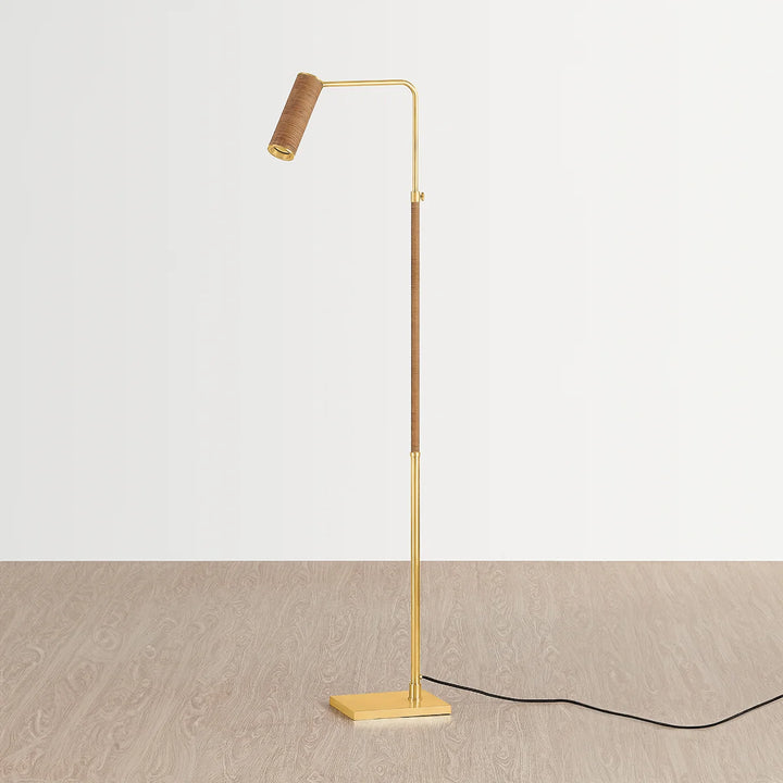 Brass floor reading lamp with woven rattan-wrapped stem and adjustable spotlight head on square base, shown on wood floor with cord