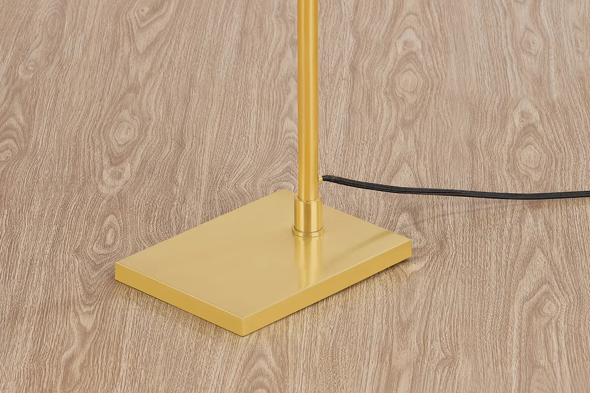 Close-up of brass floor lamp square base with metal pole and black power cord on light wood flooring