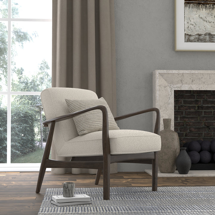 Modern upholstered accent chair with dark wood frame and throw pillow beside a fireplace in a bright living room with area rug and large window