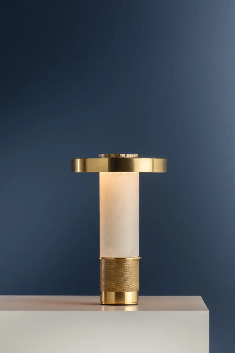 Modern brass table lamp with frosted cylindrical glass base and flat round metal shade, styled on a white pedestal against a blue wall
