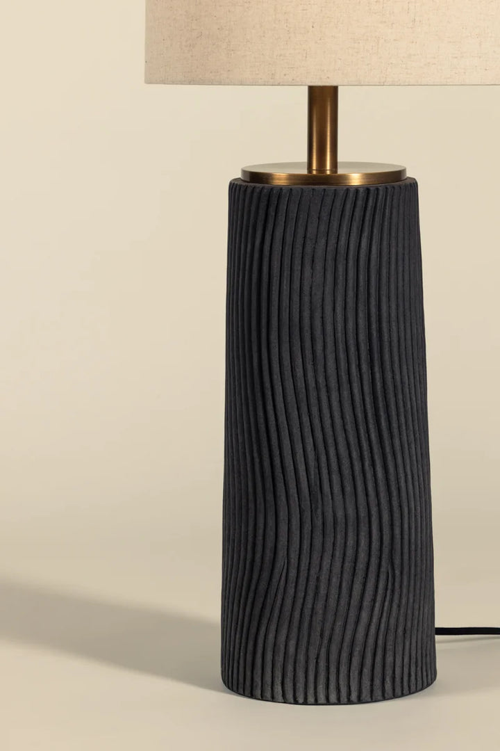 Modern table lamp with charcoal ribbed ceramic base, brass neck, and natural linen drum shade on neutral background