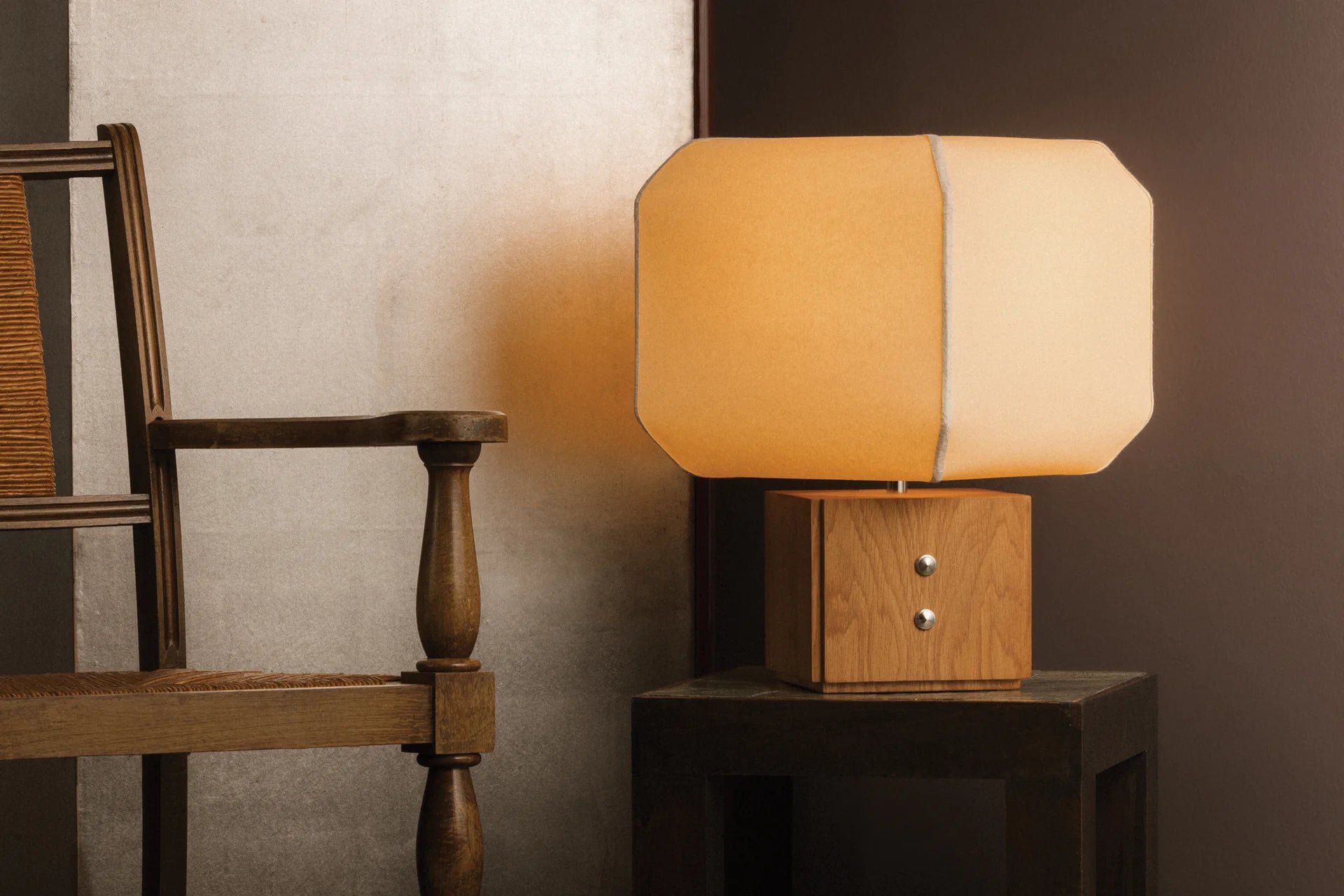 Table lamp with wooden base and beige shade on a dark surface next to a wooden chair.