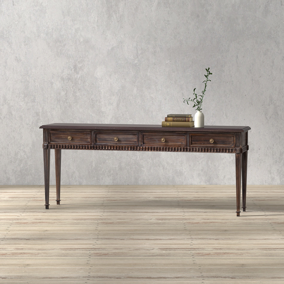A brown, rectangular Dennis Console Table with multiple drawers, placed on a wooden floor against a plain wall, featuring a decorative plant and book on top.