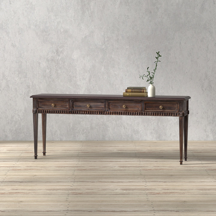 A brown, rectangular Dennis Console Table with multiple drawers, placed on a wooden floor against a plain wall, featuring a decorative plant and book on top.