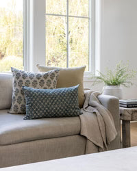 Living room with a beige sofa adorned with patterned cushions, a small table with a plant, and a window in the background.