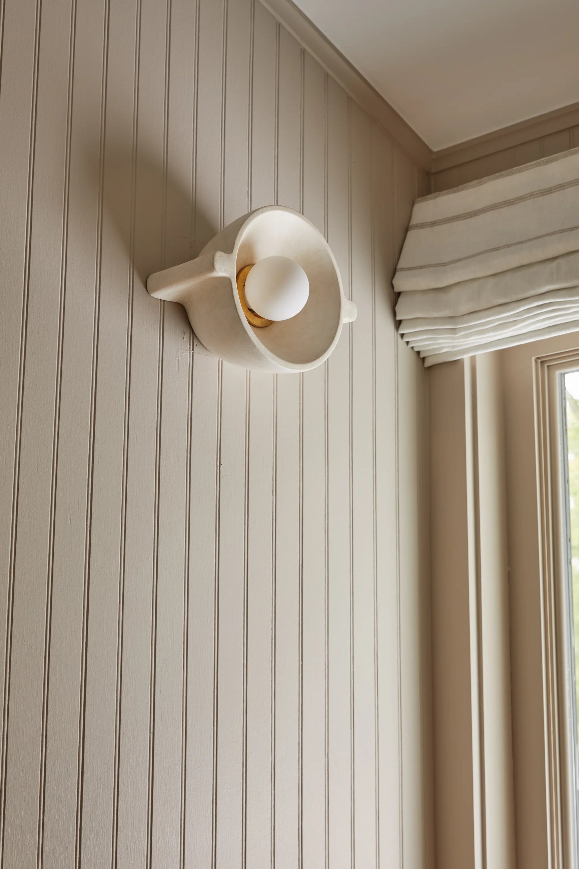 Wall-mounted light fixture on a beige paneled wall with a window in the background.