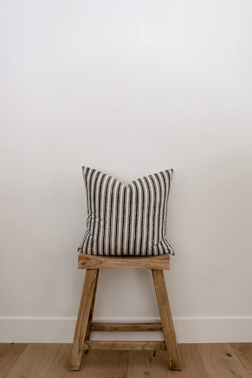 A wooden stool with a Myer Stripe Pillow Cover resting on it, adding a pop of color and comfort to the setting.