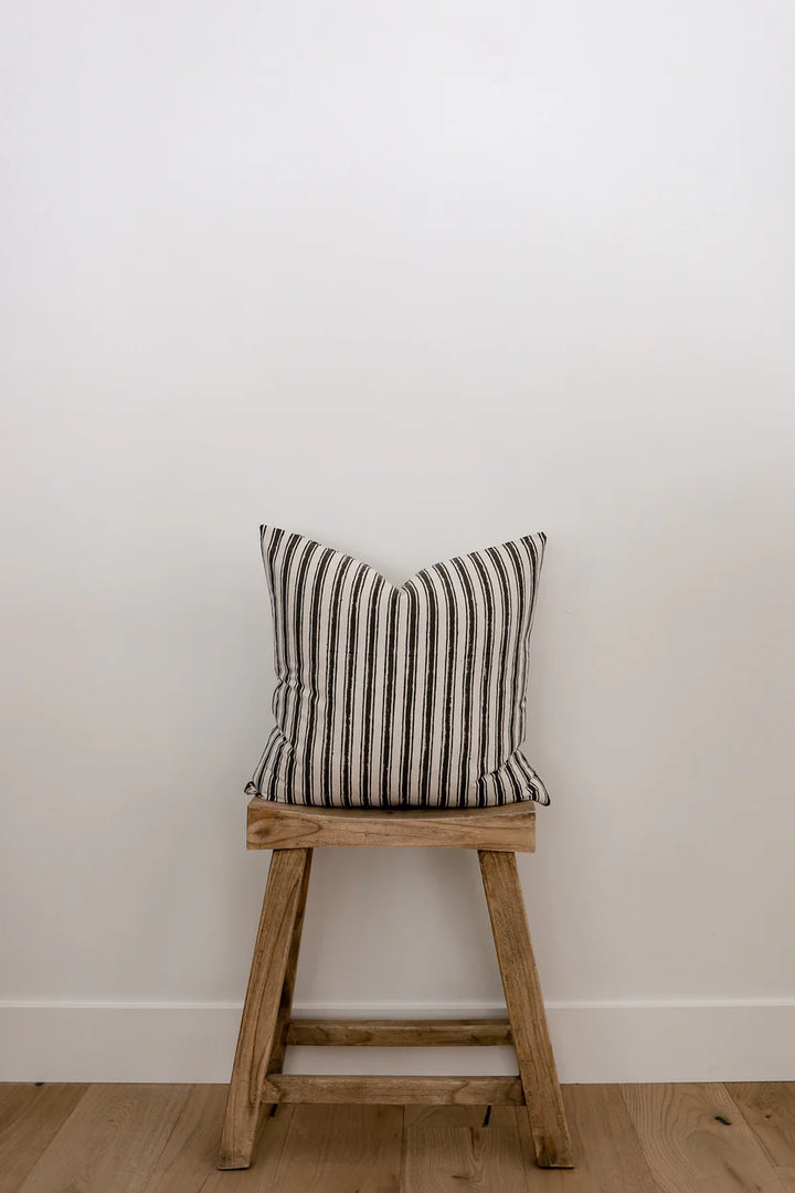 A wooden stool with a Myer Stripe Pillow Cover resting on it, adding a pop of color and comfort to the setting.