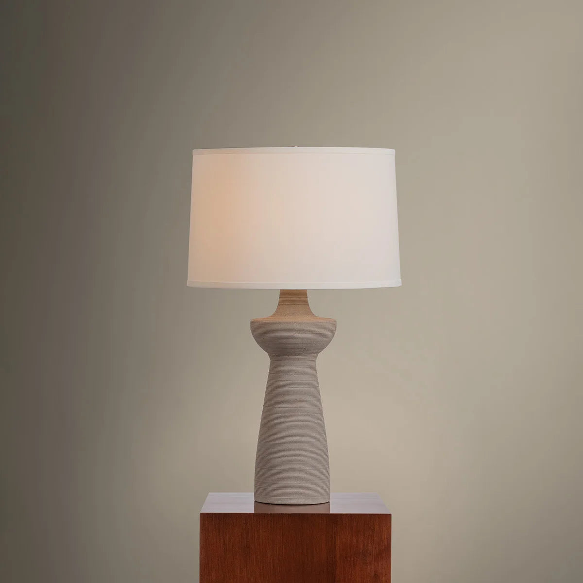 Table lamp with a beige ceramic base and white shade on a wooden pedestal against a plain wall.