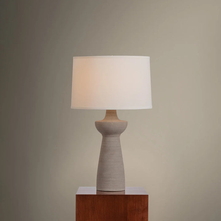 Table lamp with a beige ceramic base and white shade on a wooden pedestal against a plain wall.